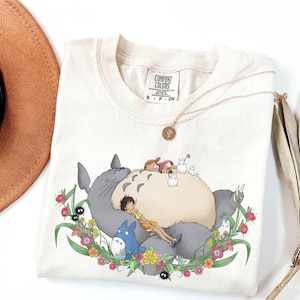 Puede incluir: Camiseta color crema con una colorida ilustraci&oacute;n de personajes de la pel&iacute;cula de animaci&oacute;n, Mi Vecino Totoro. El dise&ntilde;o incluye a Totoro, Mei y otros esp&iacute;ritus del bosque rodeados de flores. Un collar dorado descansa sobre la camisa.