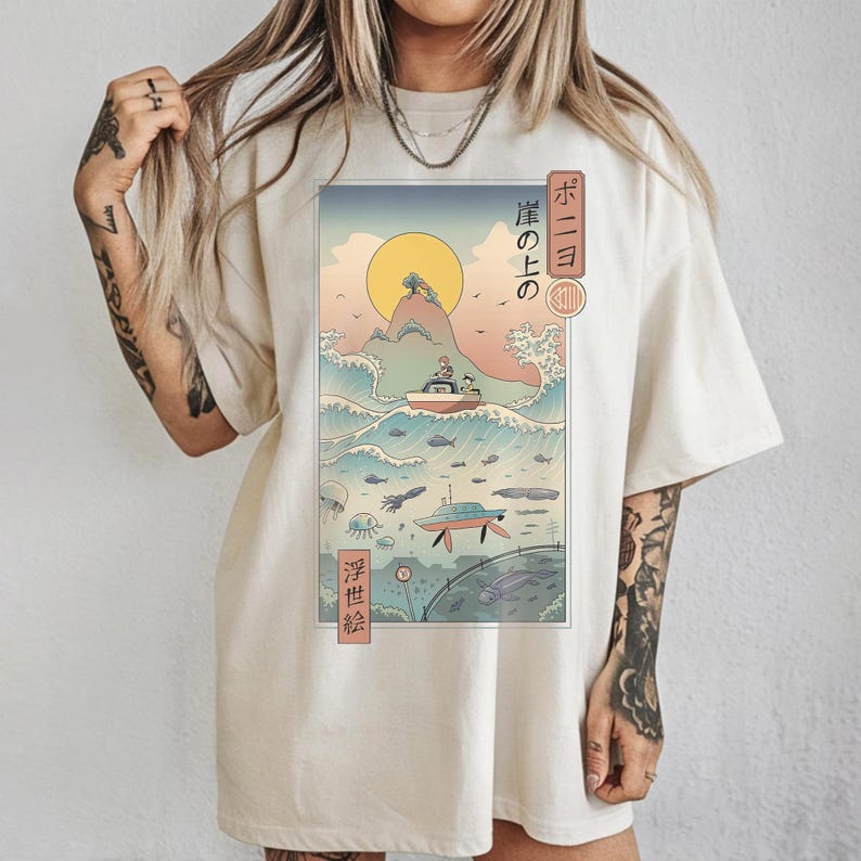 May include: Cream-colored t-shirt featuring a Japanese-inspired graphic. The artwork depicts a boat on a wave, a setting sun, and Japanese text. The design is framed with a border and includes additional marine life illustrations.