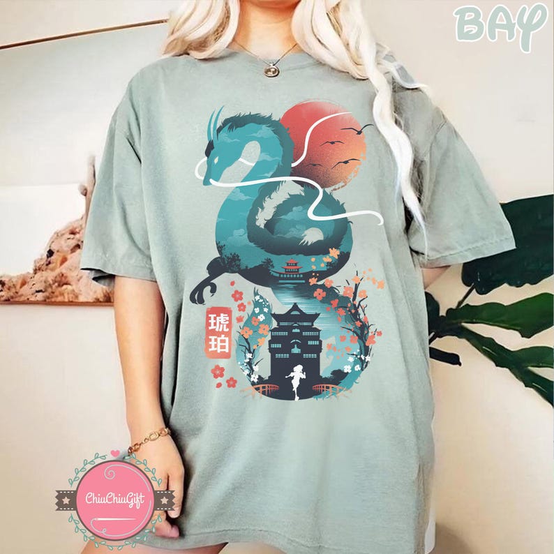 Puede incluir: Camiseta verde salvia con un dise&ntilde;o de drag&oacute;n turquesa, un sol rojo y texto japon&eacute;s. El gr&aacute;fico incluye una pagoda, flores de cerezo y la silueta de una persona. La camiseta es de corte holgado.