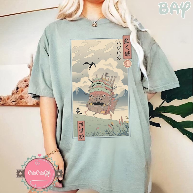 Pu&ograve; includere: T-shirt verde salvia con un'illustrazione in stile giapponese di un castello in movimento e testo giapponese. L'opera d'arte raffigura una struttura dettagliata del castello su un paesaggio con montagne, nuvole e un uccello in volo.