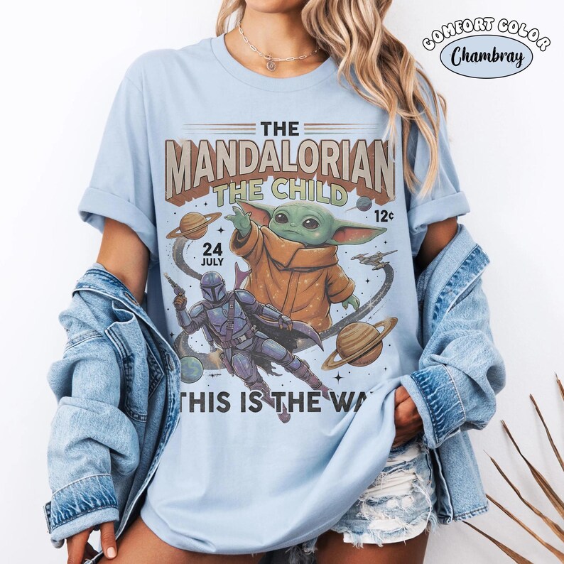 May include: A light blue t-shirt featuring a graphic print of The Mandalorian and The Child in space. The text reads THE MANDALORIAN THE CHILD 24 JULY THIS IS THE WAY. The design includes planets and a spaceship. A small circular label says COMFORT COLOR Chambray.
