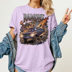 Pode incluir: Uma t-shirt roxo-claro com uma impress&atilde;o gr&aacute;fica de um carro azul estilizado com uma express&atilde;o zangada, rodeado por chamas e detritos. O texto RAMONE est&aacute; impresso numa fonte met&aacute;lica acima do carro em movimento.