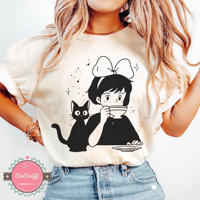 Pu&ograve; includere: T-shirt color crema con un'illustrazione in bianco e nero di una ragazza con un fiocco, che beve da una tazza, e un gatto nero. Il design include stelle e il testo "ChiuChiuGift" su un logo rosa e verde.