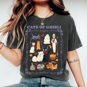 K&ouml;nnte beinhalten: Dunkelgraues T-Shirt mit einer Grafik von Katzen aus Studio Ghibli-Filmen, darunter Jiji und Totoro. Das Design ist gerahmt und enth&auml;lt den Text "CATS OF GHIBLI". Das T-Shirt ist bequem und f&uuml;r den Alltag geeignet.