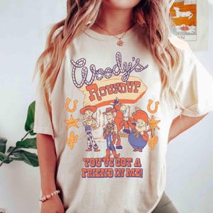 May include: Cream-colored t-shirt featuring the text "Woody's Roundup" in a stylized font, with cartoon characters and the phrase "You've Got a Friend in Me!" in orange and purple. The design includes horseshoes, stars, and cacti.