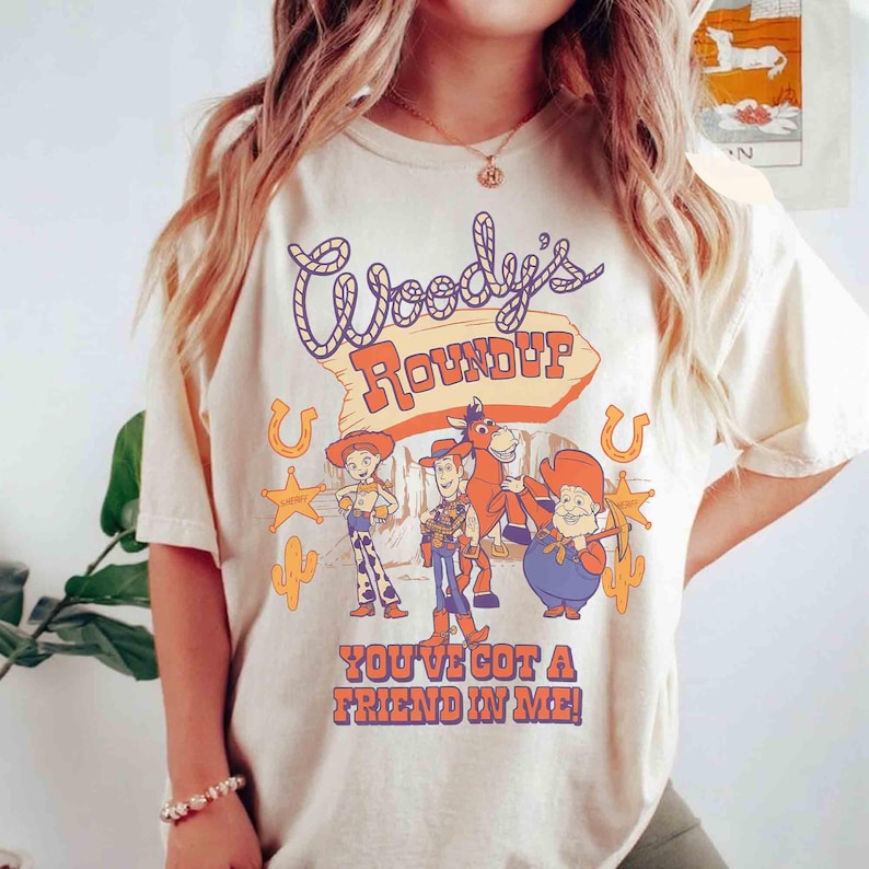 May include: Cream-colored t-shirt featuring the text "Woody's Roundup" in a stylized font, with cartoon characters and the phrase "You've Got a Friend in Me!" in orange and purple. The design includes horseshoes, stars, and cacti.