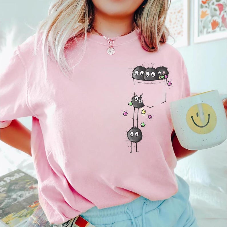 May include: A light pink sweatshirt featuring a pocket with a cartoon design of black, fluffy creatures with multiple eyes. The design includes colorful star accents and a smiling face mug.