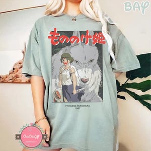 May include: A pale green t-shirt with a graphic from the animated film Princess Mononoke. The graphic features the Japanese title in red, a character illustration, and the text "PRINCESS MONONOKE 1997". The shirt has short sleeves and a loose fit.