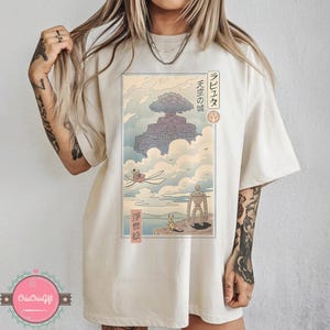May include: Cream-colored t-shirt featuring a Japanese anime-style illustration. The artwork depicts a floating castle, characters, and Japanese text. The design is in shades of blue, gray, and beige.