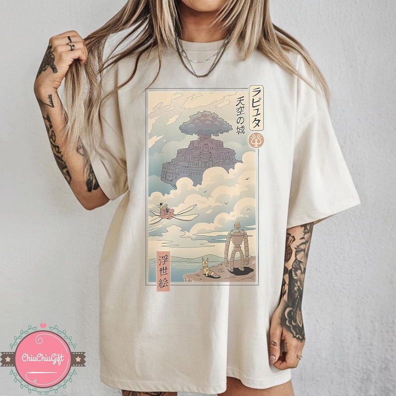 May include: Cream-colored t-shirt featuring a Japanese anime-style illustration. The artwork depicts a floating castle, characters, and Japanese text. The design is in shades of blue, gray, and beige.