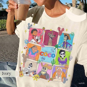 May include: Ivory-colored t-shirt featuring a colorful graphic of characters from the Disney Pixar movie "Coco." The design includes various characters in a grid-like arrangement, with the movie's title prominently displayed. The shirt is made of a soft material.