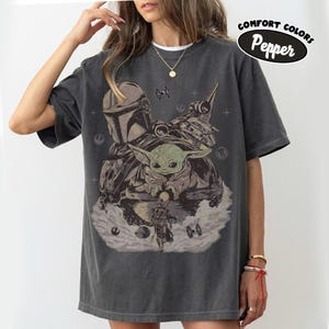 May include: A charcoal gray short-sleeve t-shirt featuring a graphic print of a helmeted character, a small green creature in a floating pod, and a starship. The design includes small star icons and the text Comfort Colors Pepper in the corner.