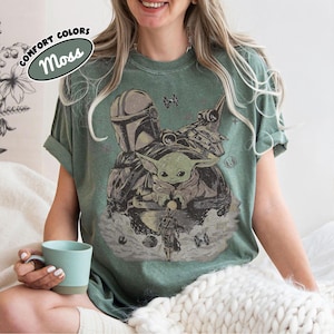 May include: A moss green crew neck t-shirt featuring a distressed graphic print of The Mandalorian and Grogu. The design includes a TIE fighter, a starship, and small celestial elements. A label in the corner reads COMFORT COLORS Moss.