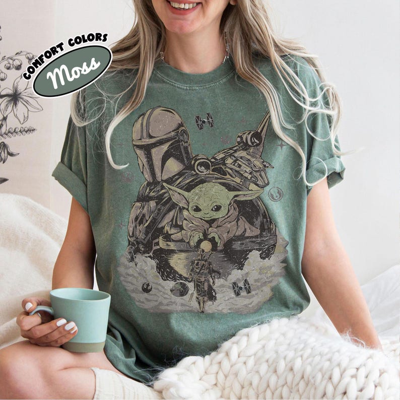 May include: A moss green crew neck t-shirt featuring a distressed graphic print of The Mandalorian and Grogu. The design includes a TIE fighter, a starship, and small celestial elements. A label in the corner reads COMFORT COLORS Moss.