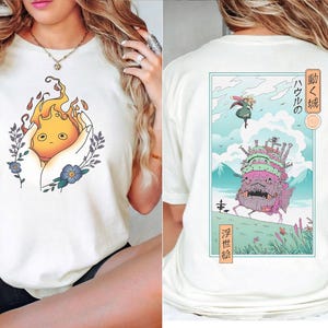 K&ouml;nnte beinhalten: Cremefarbenes T-Shirt mit Cartoon-Illustration. Auf der Vorderseite ist eine feurige Kreatur in H&auml;nden zu sehen, umgeben von floralen Akzenten. Die R&uuml;ckseite zeigt eine gerahmte Szene mit einem sich bewegenden Schloss und japanischem Text.