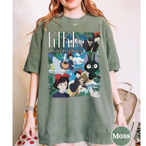 May include: Moss green t-shirt featuring a graphic from Kiki's Delivery Service. The design includes the text "Kiki's Delivery Service" and illustrations of the main characters, including Kiki and Jiji the cat. The shirt is a relaxed fit.