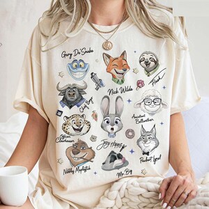 Op de afbeelding: Cr&egrave;mekleurig T-shirt met cartoonillustraties van personages uit de film Zootropolis, waaronder Nick Wilde, Judy Hopps en Flash. De namen van de personages staan boven of onder hun afbeeldingen. Het shirt heeft een ronde hals en korte mouwen.