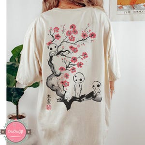 May include: Cream-colored t-shirt featuring a Japanese-inspired design. The back of the shirt displays a black and red illustration of a flowering tree with three spirit-like figures. Japanese text and a red seal are also present.