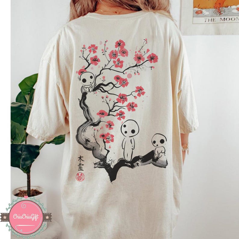 May include: Cream-colored t-shirt featuring a Japanese-inspired design. The back of the shirt displays a black and red illustration of a flowering tree with three spirit-like figures. Japanese text and a red seal are also present.