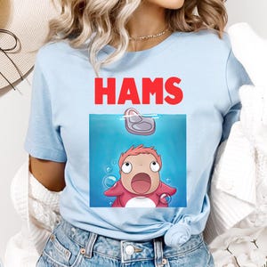 May include: Pale blue t-shirt featuring the word "HAMS" in red above a cartoon graphic. The graphic shows a character looking up at a piece of ham in water, with bubbles around it.