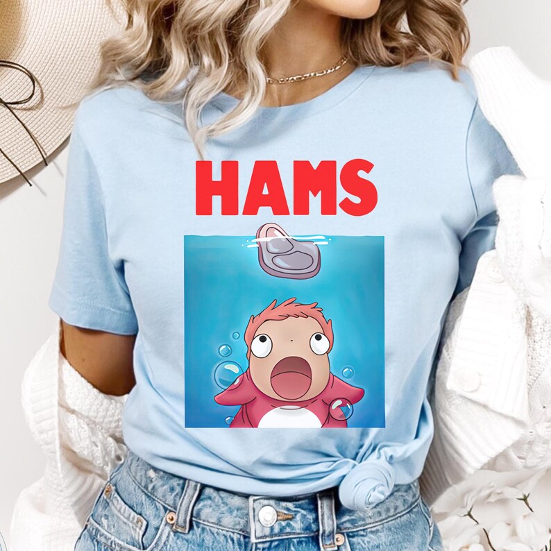 May include: Pale blue t-shirt featuring the word "HAMS" in red above a cartoon graphic. The graphic shows a character looking up at a piece of ham in water, with bubbles around it.