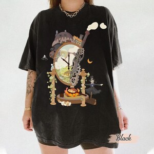 Op de afbeelding: Zwart t-shirt met een grillig klokontwerp met een cartoonfiguur, tandwielen en een halve maan. De wijzerplaat geeft de tijd aan. Het woord "Black" staat rechtsonder op de shirt.