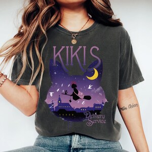 以下が含まれることがあります： 夜空のシーンが描かれた猫のシルエットのデザインのダークグレーのTシャツ。デザインには、ほうきに乗った魔女、三日月、紫色の文字で「KIKIS Delivery Service」が含まれています。