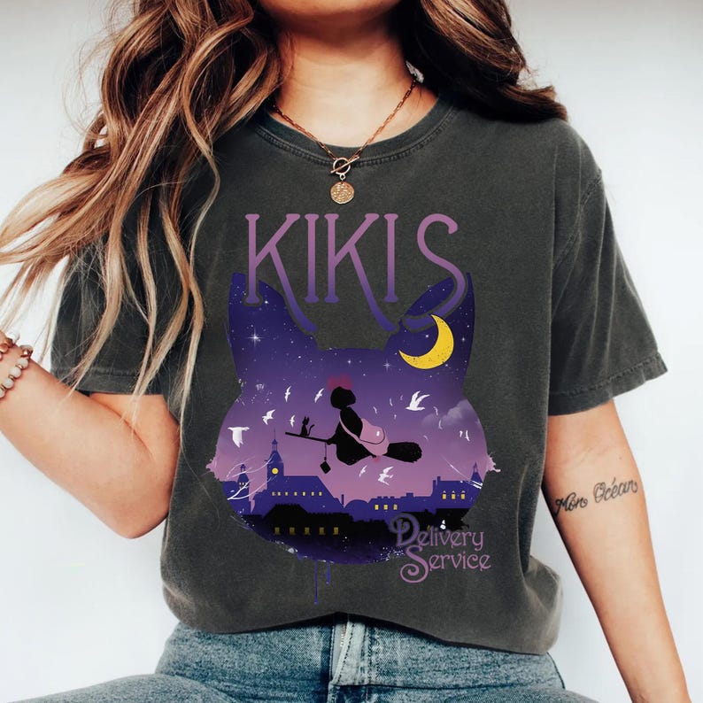 以下が含まれることがあります： 夜空のシーンが描かれた猫のシルエットのデザインのダークグレーのTシャツ。デザインには、ほうきに乗った魔女、三日月、紫色の文字で「KIKIS Delivery Service」が含まれています。