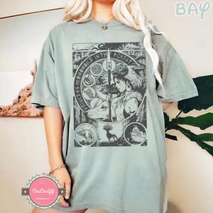 Puede incluir: Camiseta verde salvia con estampado gr&aacute;fico. El estampado presenta a una mujer con una lanza, un lobo y el texto "Princess Mononoke in the Forest". Tambi&eacute;n hay ilustraciones de animales. La camiseta tiene mangas cortas y un corte holgado.