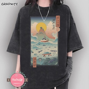 May include: Dark gray t-shirt featuring a Japanese-inspired illustration of a boat on waves, a rising sun, and marine life. The graphic includes Japanese text and a retro aesthetic. The shirt has short sleeves and a relaxed fit.