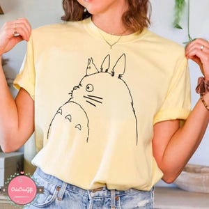 Puede incluir: Una camiseta amarilla clara con un dibujo lineal negro de un personaje de dibujos animados. El personaje tiene ojos grandes, bigotes y una peque&ntilde;a criatura en la cabeza. La camiseta es de material suave.