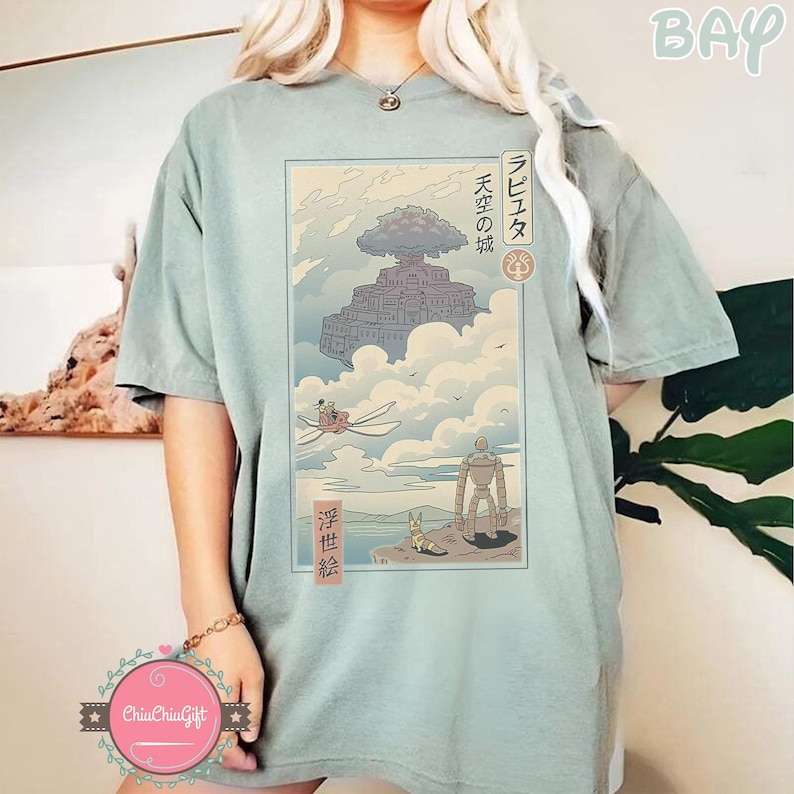 May include: Sage green t-shirt featuring a Japanese anime-style graphic. The graphic depicts a floating castle in the clouds, with Japanese text and illustrations of characters and a flying machine. The shirt has short sleeves and a relaxed fit.