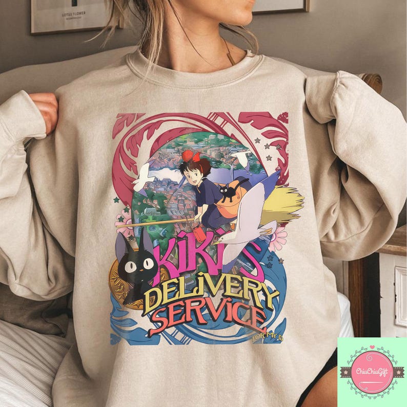 May include: Beige sweatshirt with a vibrant graphic from the animated film Kiki's Delivery Service. The design features the text "Kiki's Delivery Service" alongside illustrations of Kiki, Jiji the cat, and other characters. The sweatshirt has a crew neck and long sleeves.