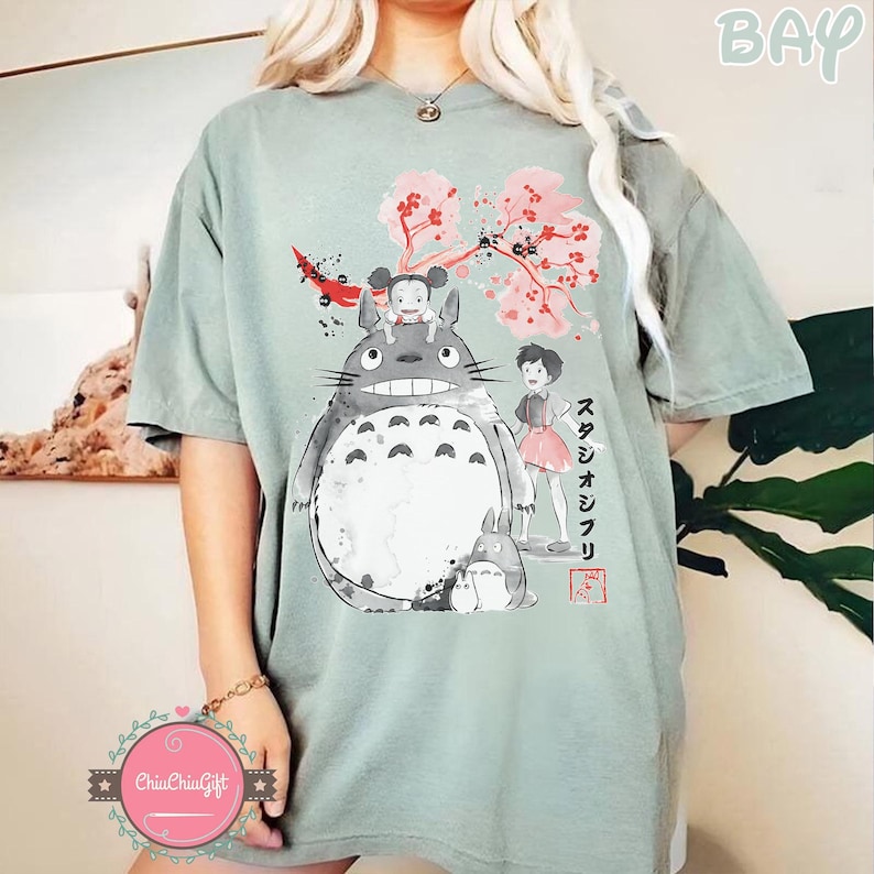 Pode incluir: T-shirt verde s&aacute;lvia com um gr&aacute;fico de Totoro e outros personagens do filme de anima&ccedil;&atilde;o Meu Vizinho Totoro. O design inclui flores de cerejeira e texto japon&ecirc;s. A camisa tem um ajuste folgado.