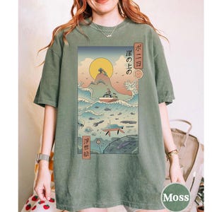 May include: Moss green t-shirt featuring a Japanese-inspired graphic. The design includes a boat on waves, a rising sun, and Japanese text. The shirt has a relaxed fit and short sleeves. The graphic is in shades of blue, yellow, and green.