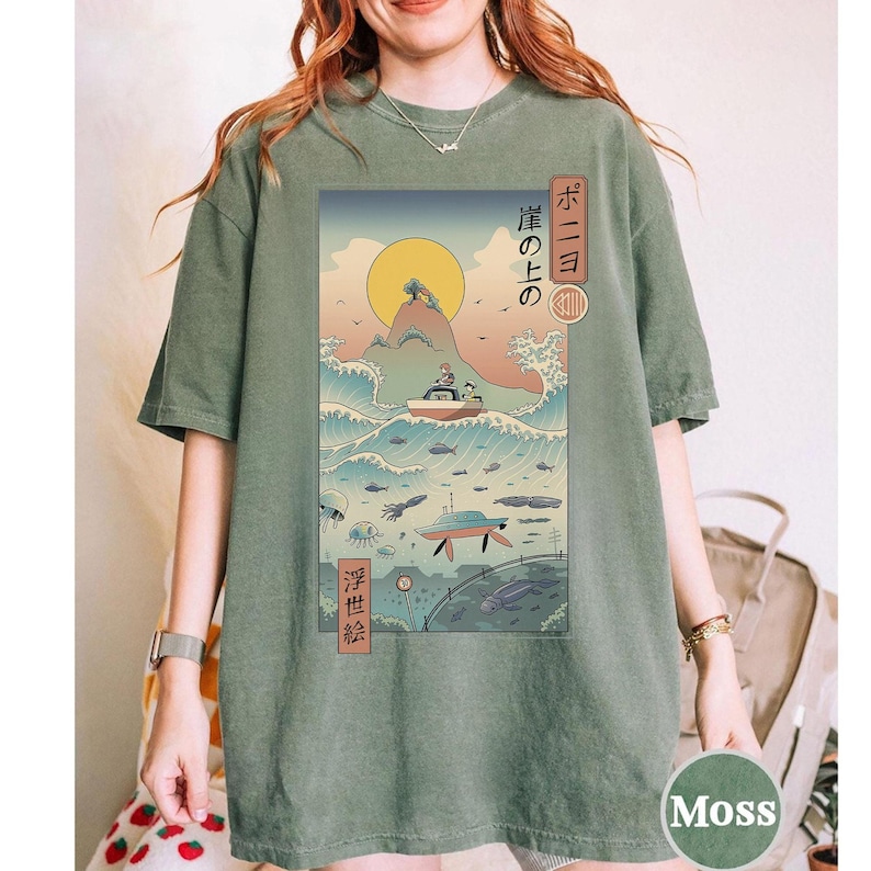 May include: Moss green t-shirt featuring a Japanese-inspired graphic. The design includes a boat on waves, a rising sun, and Japanese text. The shirt has a relaxed fit and short sleeves. The graphic is in shades of blue, yellow, and green.