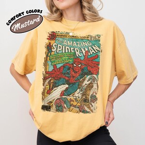 K&ouml;nnte beinhalten: Ein senfgelbes Kurzarm-T-Shirt mit einem Vintage-Marvel-Comic-Cover-Aufdruck von The Amazing Spider-Man. Der Druck zeigt Spider-Man in seinem rot-blauen Anzug mit dem Text The Amazing Spider-Man sowie dem Schriftzug Comfort Colors.