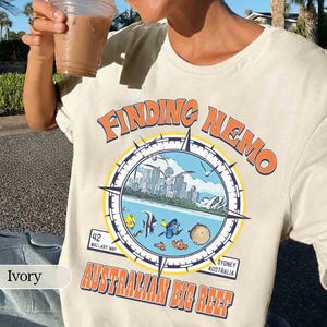 Puede incluir: Camiseta de color marfil con un diseño gráfico colorido de la película "Buscando a Nemo". El diseño incluye una brújula con peces, gaviotas y el texto "Australian Big Reef". La camiseta también tiene el texto "42 Wallaby Way, Sydney Australia."