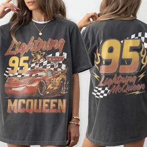 May include: A charcoal gray t-shirt featuring a graphic of the red race car Lightning McQueen with the number 95, checkered flag patterns, and the text Lightning McQueen. The back of the shirt displays a large 95 with flames and checkered accents.