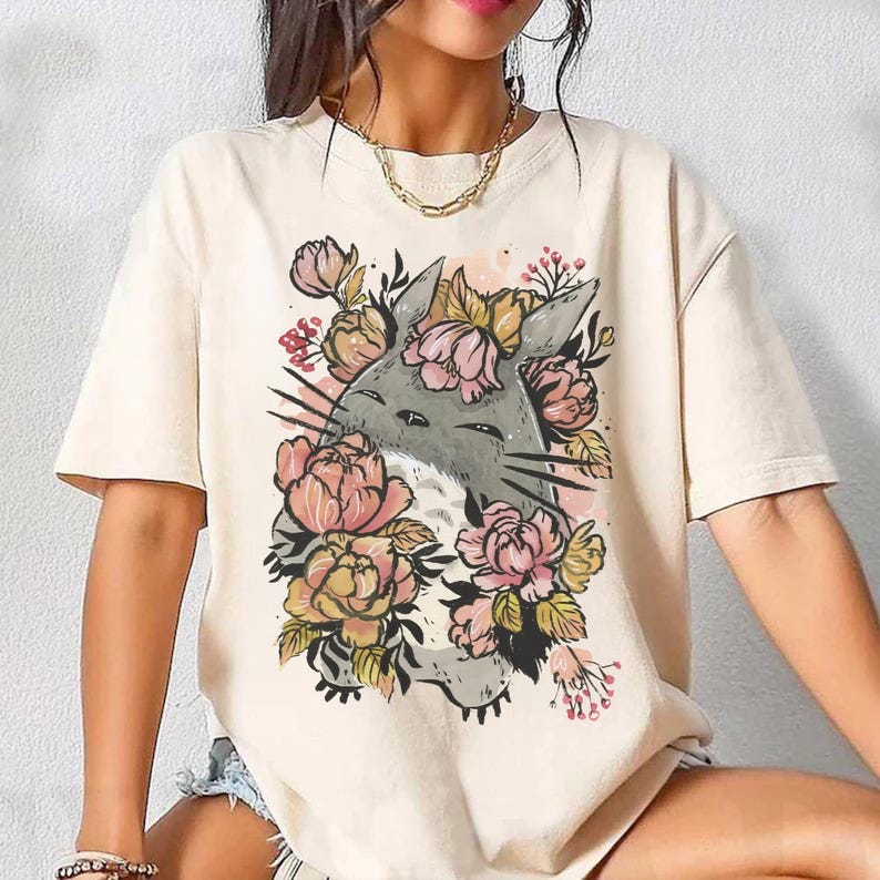 May include: Cream-colored t-shirt featuring a gray cartoon character surrounded by pink, yellow, and green floral elements. The graphic is centered on the front of the shirt. The shirt has short sleeves and a crew neck.
