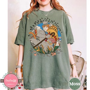 May include: Moss green t-shirt featuring a cartoon illustration of a cat in a suit with a cane, set against a backdrop of a building and bats. The design is enclosed in a blue circle. The shirt is a comfortable, casual style.