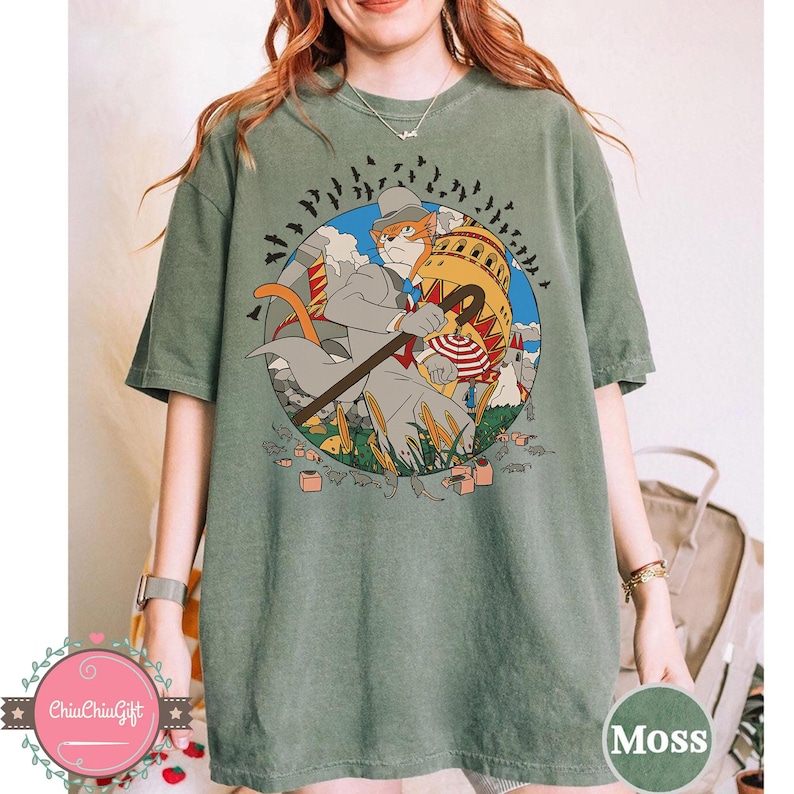 May include: Moss green t-shirt featuring a cartoon illustration of a cat in a suit with a cane, set against a backdrop of a building and bats. The design is enclosed in a blue circle. The shirt is a comfortable, casual style.