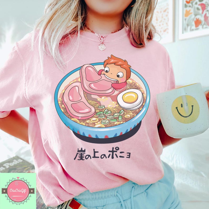 May include: Pink t-shirt featuring a cartoon ramen bowl with a character, meat slices, and an egg. The shirt has Japanese text below the bowl. A white mug with a yellow smiley face is held in the hand.