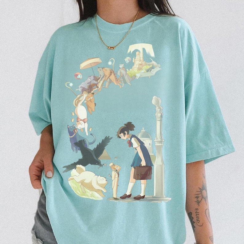 Puede incluir: Camiseta azul claro con una ilustraci&oacute;n de estilo anime. El dise&ntilde;o presenta a una colegiala, animales y elementos arquitect&oacute;nicos. La camiseta tiene un corte holgado y mangas cortas.