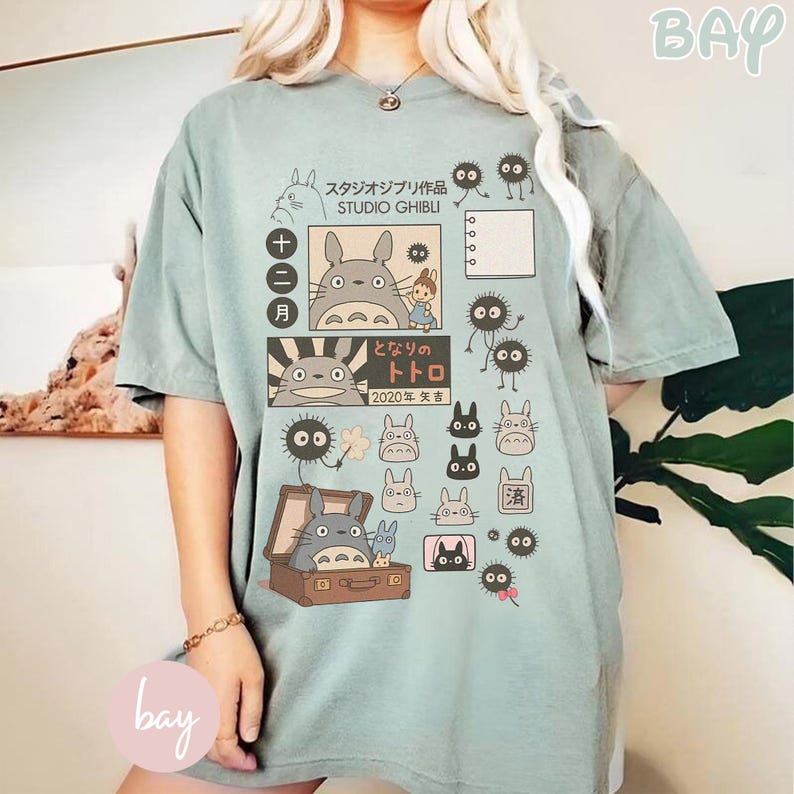 May include: A light green t-shirt featuring Studio Ghibli characters and Japanese text. The design includes illustrations of Totoro, soot sprites, and other characters from the animated film. The shirt has a relaxed fit and short sleeves.