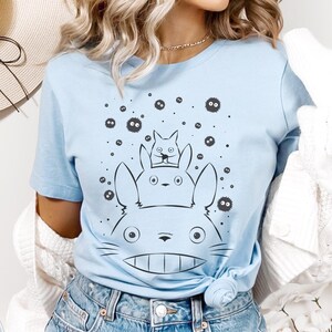 May include: Light blue t-shirt featuring a black and white graphic of three cartoon characters, with a large smiling character at the bottom and smaller characters stacked above. Small black dots are scattered around the characters.