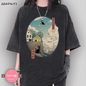 Op de afbeelding: Grafietkleurig T-shirt met een cirkelvormige illustratie van een heks op een bezem over een stad en een zwarte kat. Het ontwerp bevat Japanse tekst en afbeeldingen van huizen, vogels en wolken.
