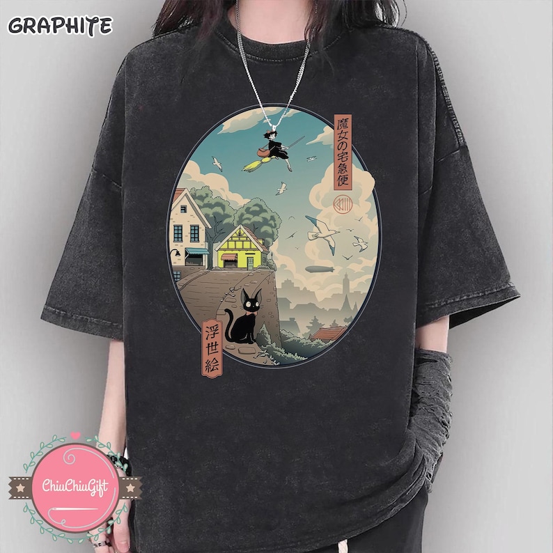 Op de afbeelding: Grafietkleurig T-shirt met een cirkelvormige illustratie van een heks op een bezem over een stad en een zwarte kat. Het ontwerp bevat Japanse tekst en afbeeldingen van huizen, vogels en wolken.