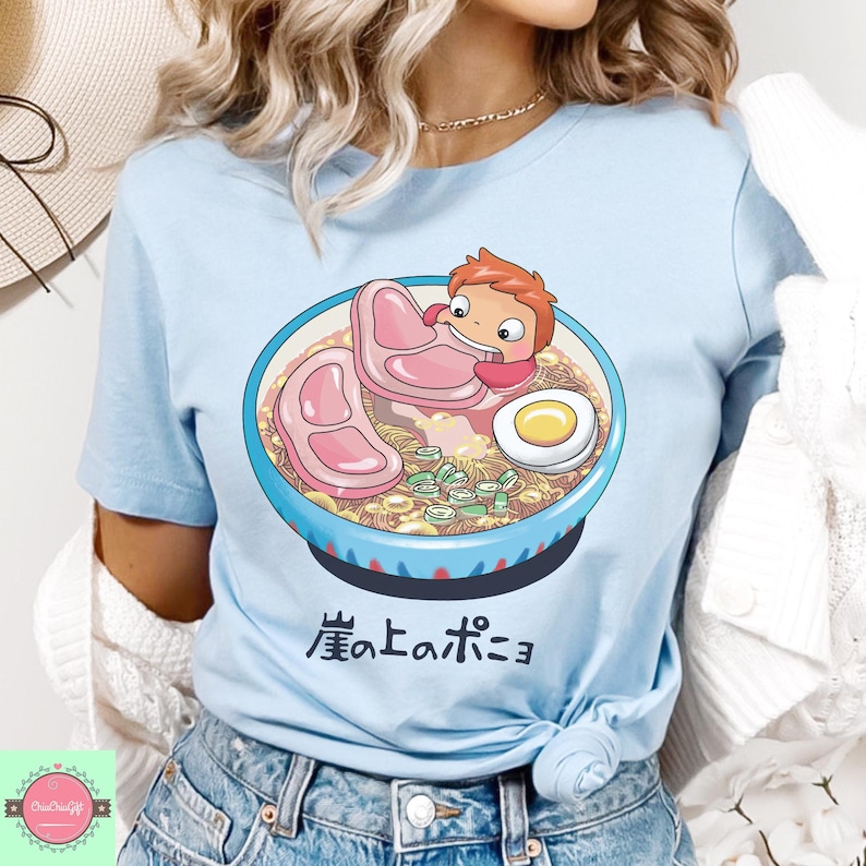 May include: Light blue t-shirt featuring a cartoon illustration of a bowl of ramen with a character holding a piece of meat. The ramen bowl is blue with a white rim and contains noodles, meat, and an egg. Japanese text is below the bowl.