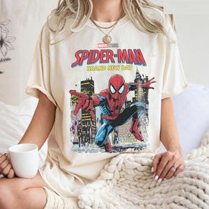May include: Cream-colored t-shirt featuring a Marvel Studios Spider-Man graphic. The graphic shows Spider-Man in a dynamic pose against a cityscape, with the words "Spider-Man Brand New Day" in red and yellow.